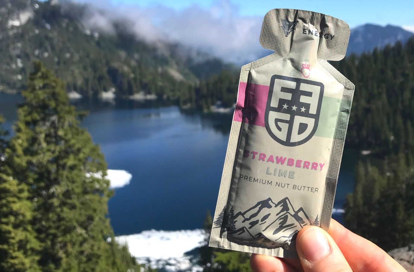 hand holding up a fat fit go premium nut butter in front of a wilderness scene